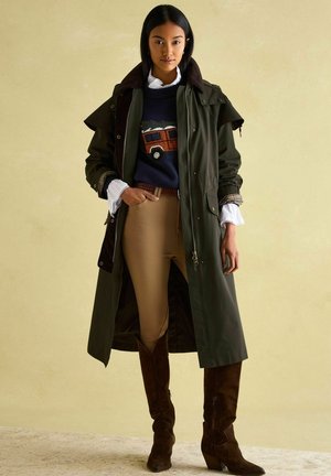 Olivengrüner langer Mantel mit einem braunen Kragen, getragen über einem marineblauen Pullover mit einem roten Fahrzeug. Beige enge Hose und braune kniehohe Stiefel.