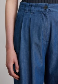 Blue denim trousers with a relaxed fit, featuring front pleats, a button closure, and side pockets. Smooth texture with a slightly faded finish.