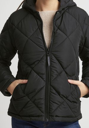 Vrouw draagt een zwarte doorgestikte jas met een rits aan de voorkant, capuchon en handen in de zakken, over een beige trui.