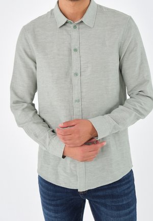 Uomo che indossa una camicia a maniche lunghe con bottoni di un verde chiaro e jeans blu, mani incrociate in vita, su uno sfondo bianco.