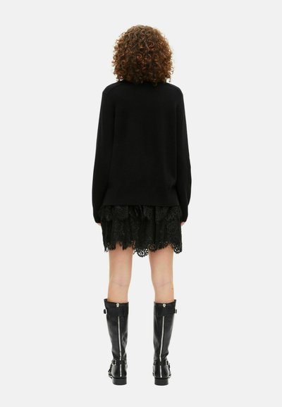 Persona con cabello rizado, vistiendo un suéter negro, una falda de encaje negro y botas altas negras con cremalleras plateadas, de pie de espaldas.