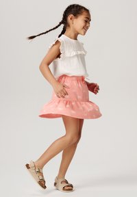 Mädchen in einem weißen, strukturierten Oberteil mit voluminösen Ärmeln und einem rosa Rock mit ausgefallenem Saum und dezentem Blumenmuster, trägt beigefarbene Sandalen.