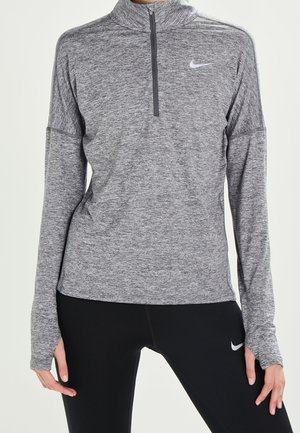 Frau trägt ein graues Nike Langarm-Oberteil mit Reißverschluss und schwarze Nike Leggings, steht mit entspannt an den Seiten hängenden Händen.