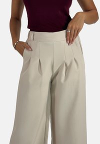 Beige wijde broek met plooien, op maat gemaakte tailleband en zijzakken. Glad stoftextuur met minimale details.