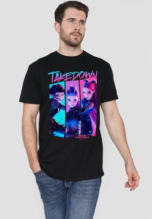 Man wearing black T-shirt featuring three animated female characters with "TAKEDOWN" text and "KPOP Demon Hunters" logo.