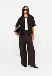 Brown knit cardigan with short sleeves and a button front, paired with dark, wide-leg trousers. Black sunglasses and a large black bag.