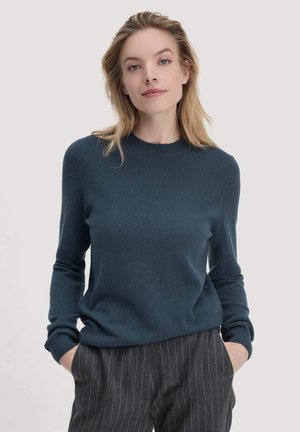 Dunkelblauer Pullover mit einem gerippten Rundhals-Ausschnitt und Bündchen, aus weichem Material. Das Modell trägt ihn zu grauen Nadelstreifen-Hosen.