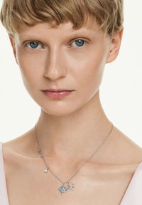 Kvinna med kort blont hår som bär ett silverhalsband med två stjärnformade hängen och en liten klar stencharm.