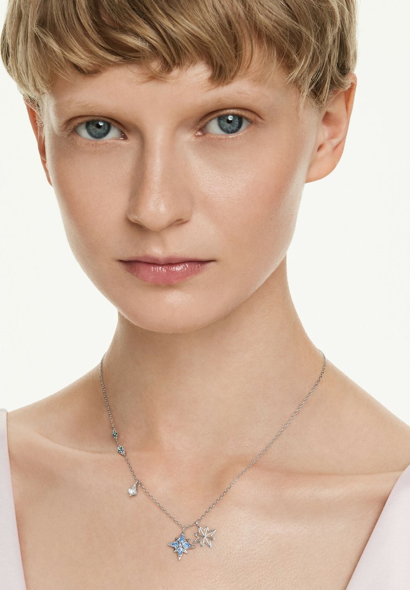 Kvinna med kort blont hår som bär ett silverhalsband med två stjärnformade hängen och en liten klar stencharm.