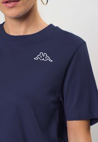 Ženska nosi temno modro majico s kratkimi rokavi z majhnim belim logotipom dveh oseb, ki sedita nasproti ena drugi, na levi strani prsi.