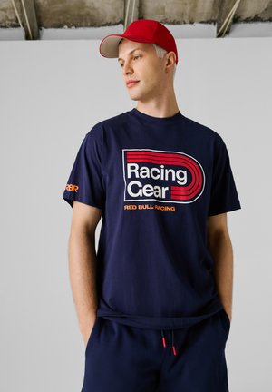 Moški v rdeči kapici in temno modri majici z napisom »Racing Gear Red Bull Racing«, stoji z rokami v žepih na enobarvnem ozadju.