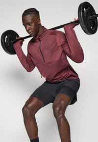 Maroon sportska jakna sa rajsferšlusom, geometrijski uzorak; crne kratke hlače; osoba podiže uteg. Fokus na fitness položaj i detalje odjeće.