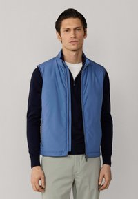 Mladý muž v modré vesta bez rukávů se zipem přes černé tričko s dlouhým rukávem a světle šedé kalhoty, stojící před jednobarevným pozadím.