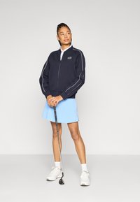 Námornícka bunda na zips s bielymi pruhmi, ľahká látka, kombinovaná so svetlomodrou sukňou. Doplnky zahŕňajú golfovú palicu a biele tenisky.