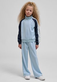 Noor juustega noor tüdruk, kes kannab helesinist ja tumesinist sportlikku dressipluusi ning valgeid tossusid, seisab tavalise tausta ees.