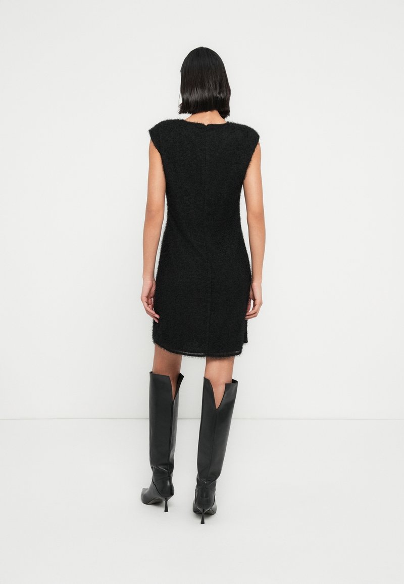 Femme avec une coupe au carré noire portant une robe noire texturée sans manches et des bottes noires à talons hautes jusqu’aux genoux, debout contre un mur blanc uni.