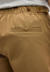 Pantalon couleur sable avec taille élastique et deux boutons marron, présentant une poche passepoilée arrière unique et un logo en relief subtil au-dessus de la poche.