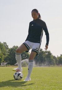 Svart langermet treningsgenser med hvite striper, hvite shorts og knelange hvite sokker. Balanserer en svart og hvit fotball på foten.