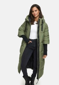 Lång olivgrön puffjacka med huva, som har en framsida med dragkedja, sidofickor och svarta detaljer. Bärs över en vit topp och svarta jeans.