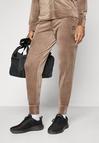Sametový teplákový komplet v svetlohnedej farbe s bundou na zips a detailom loga. Čierne tenisky s vrchnou časťou z mesh materiálu. Čierna športová taška držaná v ľavej ruke.
