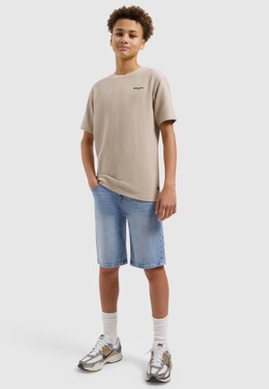 Tienerjongen staat met handen in de zakken, draagt een beige geribd T-shirt, lichtblauwe denim short, witte sokken en witte sneakers.