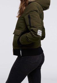 Veste rembourrée vert olive avec capuche, présentant des poches latérales zippées et un logo sur la manche. Les poignets et l'ourlet ribbed offrent de la structure.