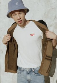 Gekleed in een wit T-shirt met een rood Levi's-logo, een bruine textuur cardigan en blauwe spijkerbroeken, aangevuld met een lichtblauwe bucket hat.