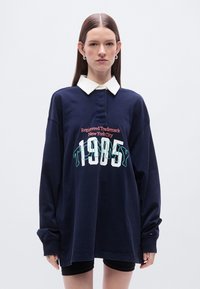 Μπλε ναυτικό πόλο πουκάμισο με λευκό γιακά, με κεντημένο κείμενο «Tommy» και «Registered Trademark New York City» σε αντίθετα χρώματα.