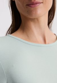 Hellgrünes, geripptes Oberteil mit weitem Ausschnitt, glatter Textur und tailliertem Design, das einen Teil des Halses und des Gesichts zeigt.