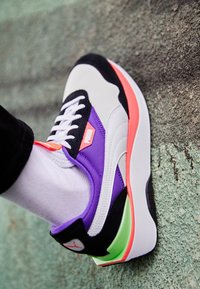 Buty sportowe Puma mają biały wierzch z czarnymi akcentami, fioletowe boczne panele oraz wyrazistą różowo-zieloną podeszwę z teksturowanym wzorem.