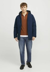 Veste matelassée bleu marine avec capuche, fermeture éclair à l'avant, portée sur un pull couleur rouille et une chemise blanche, assortie à un jean bleu et des chaussures noires.