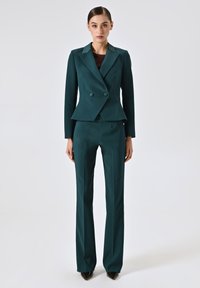 Completo personalizzato color teal con giacca doppiopetto, dotata di due bottoni e pantaloni dritti. Tessuto liscio, design strutturato e silhouette aderente.