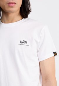 Weißes Baumwoll-T-Shirt mit einem kleinen schwarzen Logo auf der Brust und einem schwarzen Etikett mit einem orangen Design am Ärmel, kurzärmlig.