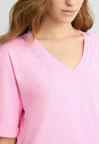 Top a manica corta con scollo a V in morbido tessuto rosa pallido. Presenta delicati accenti di strass lungo il collo. Tessuto liscio, vestibilità comoda.
