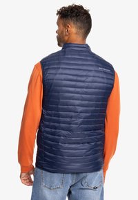 Chaleco acolchado azul marino con collar alto y material ligero, con un sutil logo en la parte posterior. Se lleva sobre una camiseta de manga larga naranja.