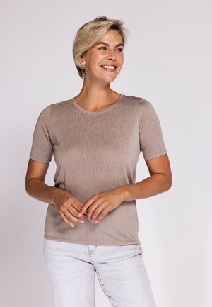 Smiling woman with short blonde hair wearing a light brown short-sleeve shirt and light-colored jeans standing against a plain background.