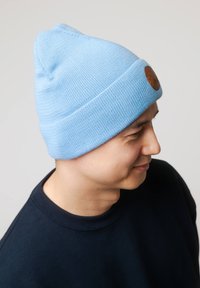 Jasnoniebieska dzianinowa czapka z nausznikami i brązową skórzaną naszywką z boku. Gładka faktura, stożkowaty kształt, odpowiednia do codziennego noszenia.