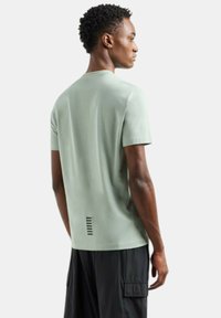 Jasnozielona koszulka z krótkim rękawem, wykonana z oddychającego materiału, z prostym krojem z tyłu i czarnym paskiem na dole.