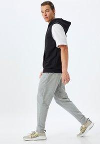 Sudadera sin mangas negra con cremallera frontal, combinada con pantalones de chándal gris claro. El modelo lleva zapatillas gris y beige con detalles de cordones.