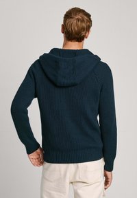 Jūrų mėlynos spalvos ganga megzta su dideliu gobtuvu, prigludusiomis rankovėmis ir gumbuotu apačia, rodoma iš nugaros prieš neutralų foną.