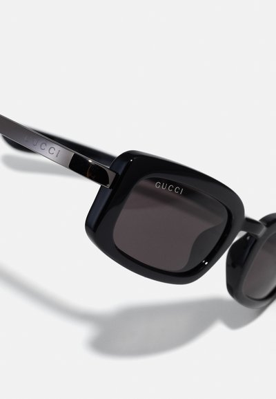 Lunettes de soleil noires avec une monture en forme de carré. Fabriquées en plastique brillant, avec des verres foncés et un logo Gucci argenté sur la branche.