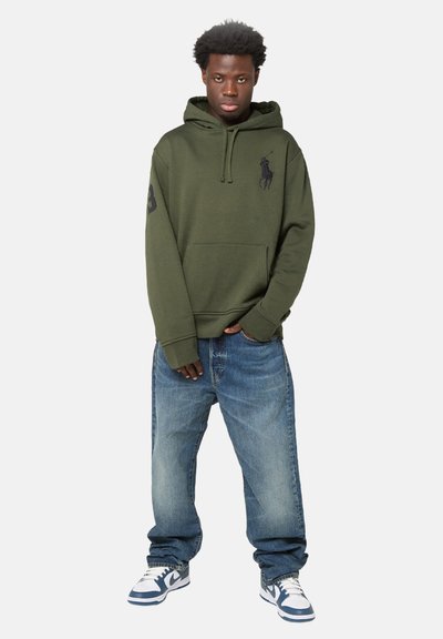 Felpa con cappuccio verde oliva con tasca a marsupio e logo; jeans blu a vestibilità comoda; sneakers bianche e blu navy con dettagli neri.