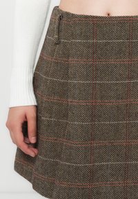 Brown herringbone-patterned skirt with red and blue accents, pleated design, at waist level, accompanied by a white knitted sleeve.