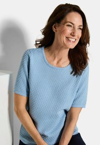 Vrouw met bruin haar, gekleed in een textuur lichtblauwe korte mouw top, lachend en kijkend naar de zijkant tegen een eenvoudige witte achtergrond.
