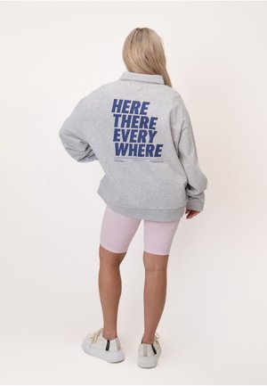 Person med langt blondt hår iført en grå sweatshirt med "HERE THERE EVERY WHERE" i blå bogstaver, lyserøde shorts og hvide sneakers.