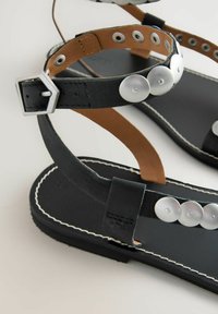 Mustad mustad mustade taldadega mustad nahksandaalid, millel on hõbedase tooni ringikujulised nupud, üks pahkluuse rakmed ja pruunist nahast detailid.