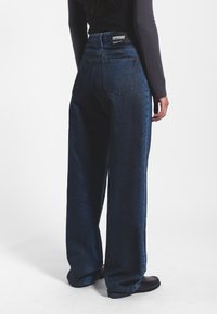 Jeans de mezclilla de pierna ancha en denim azul oscuro con cintura alta, que presentan una etiqueta negra en la parte posterior y dos bolsillos traseros de parche. Textura suave.