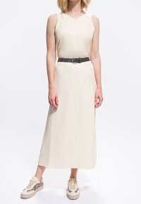 Femeie îmbrăcată într-o rochie midi crem fără mâneci, cu o curea neagră cu modele și pantofi sport asortați, stând în fața unui fundal alb.