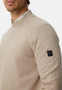 Beige knit sweater with a textured finish, ribbed crew neckline, and a black logo patch on the left sleeve. Layered over a plaid shirt.