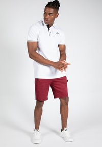 Witte polo met zwarte accenten, gecombineerd met rode sportieve shorts. De outfit heeft een moderne pasvorm en wordt aangevuld met witte sneakers.